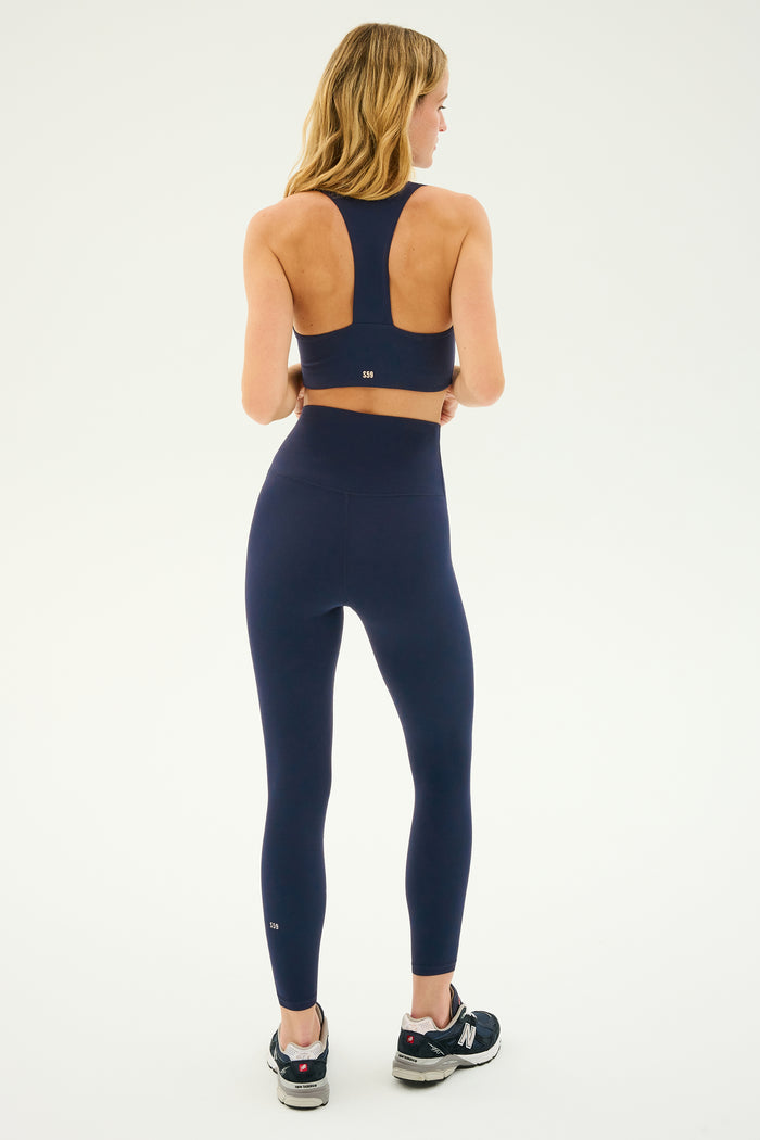 A woman with shoulder-length blonde hair is shown from the back, wearing a navy sports bra and the Airweight Super High Waist 7/8 leggings in Indigo by SPLITS59, along with black running shoes. Perfect for hot yoga, the background is plain white.