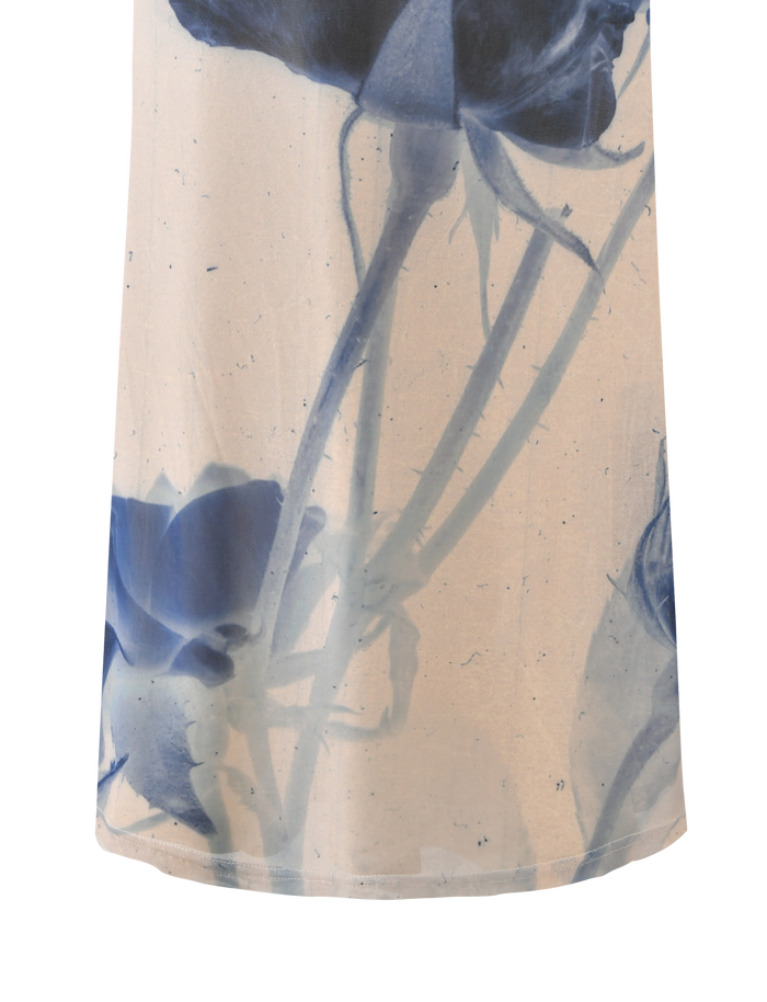 A close-up of the TIGERMIST JANIS MAXI SKIRT - INDIGO FLORAL shows semi-transparent fabric with large blue floral designs and stems on a light beige background.