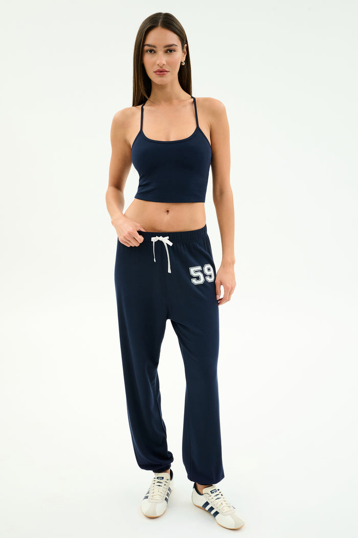 A woman stands against a plain background wearing the SPLITS59 Airweight Crop - Indigo with matching navy joggers featuring "59" on the thigh and white sneakers—a stylish look for hot yoga or casual outings.