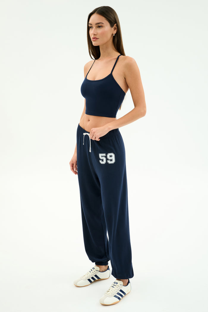 A woman wears the SPLITS59 Airweight Crop - Indigo with matching drawstring sweatpants featuring “59” on the left leg, plus white sneakers with navy stripes—ideal for hot yoga or casual outings.