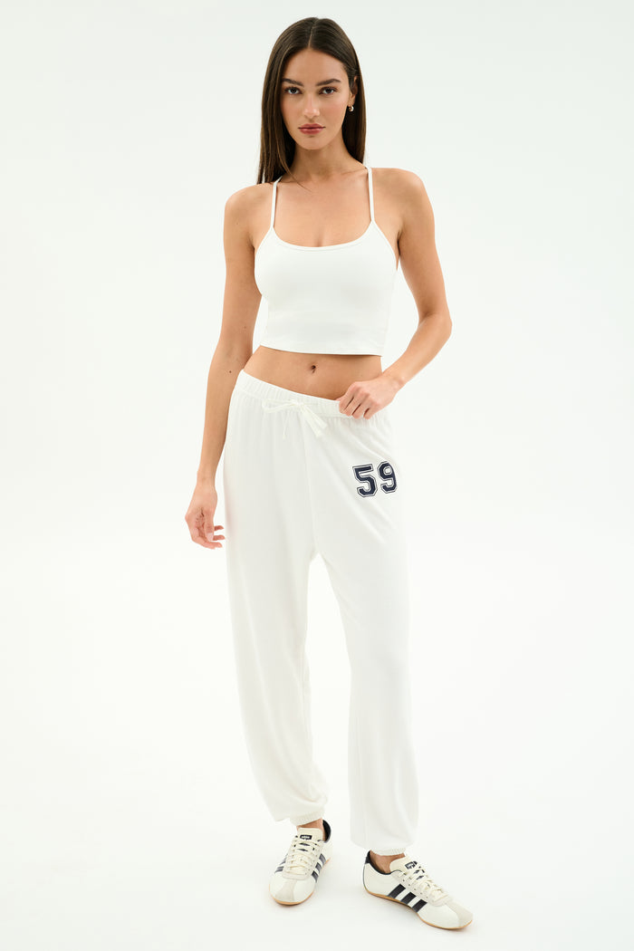 A woman wears the SPLITS59 Airweight Crop - White with white sweatpants featuring 59 and white Adidas sneakers, standing against a plain white background.