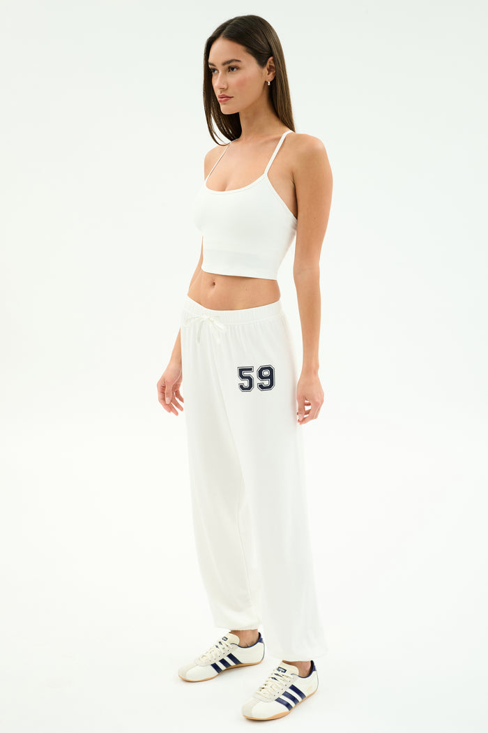 A woman poses in a white studio wearing the SPLITS59 Airweight Crop - White, matching drawstring pants with "59" on the left leg, and white sneakers with navy stripes—ideal for hot yoga or a casual outing.