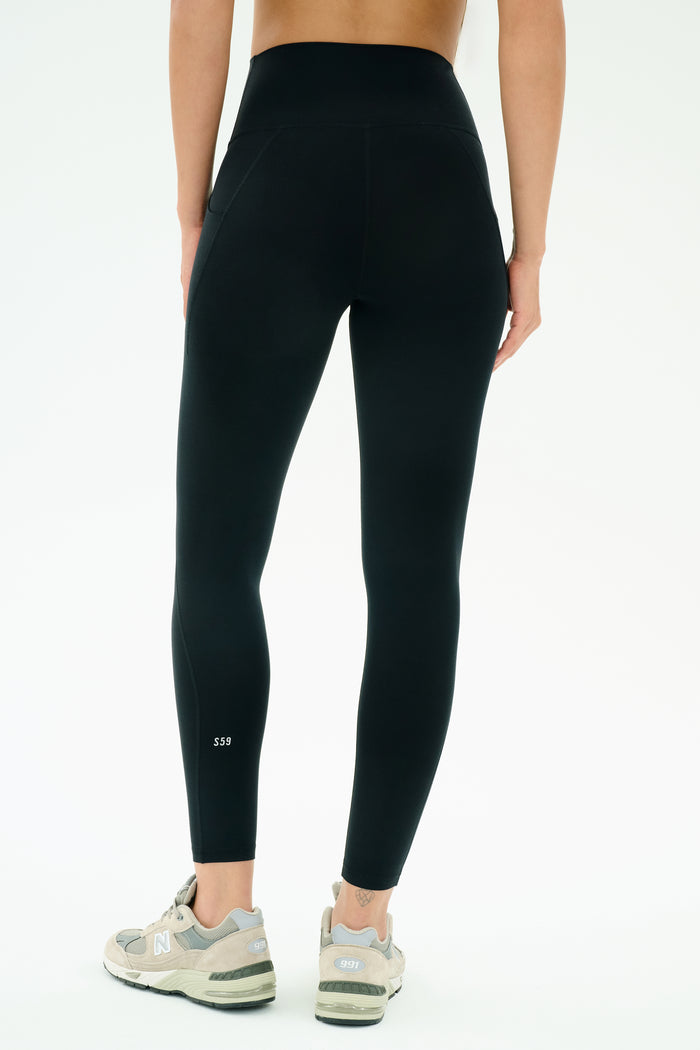 A person is shown from the back in SPLITS59 Airweight High Waist 7/8 Pocket Legging in black and grey New Balance sneakers, standing against a plain white background.
