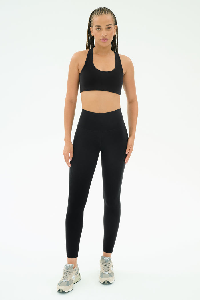 A person faces forward in SPLITS59's Airweight High Waist 7/8 Pocket Legging - Black, a black sports bra, and gray athletic shoes, pictured against a plain white background.