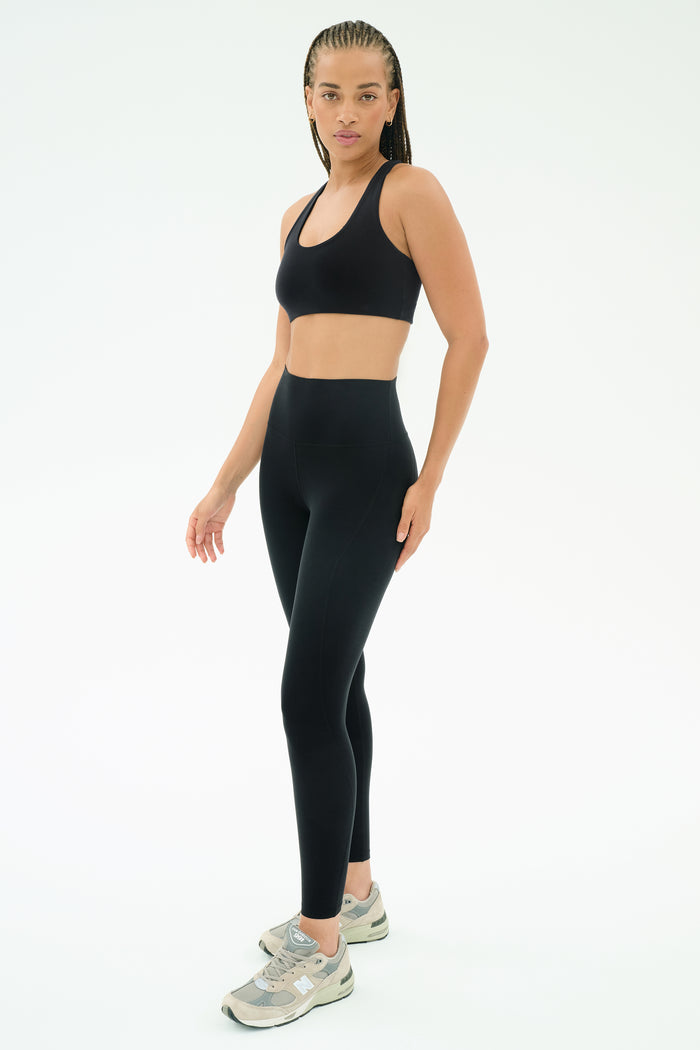 A woman wears the SPLITS59 Airweight High Waist 7/8 Pocket Legging in black, a matching sports bra, and gray sneakers, standing against a plain white background.