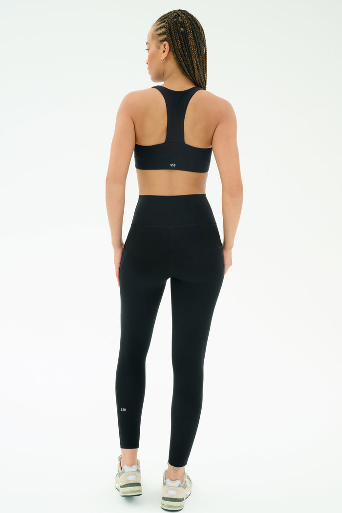 A woman with braided hair wears the SPLITS59 Airweight High Waist 7/8 Pocket Legging in black, paired with a matching sports bra, standing against a plain white background.