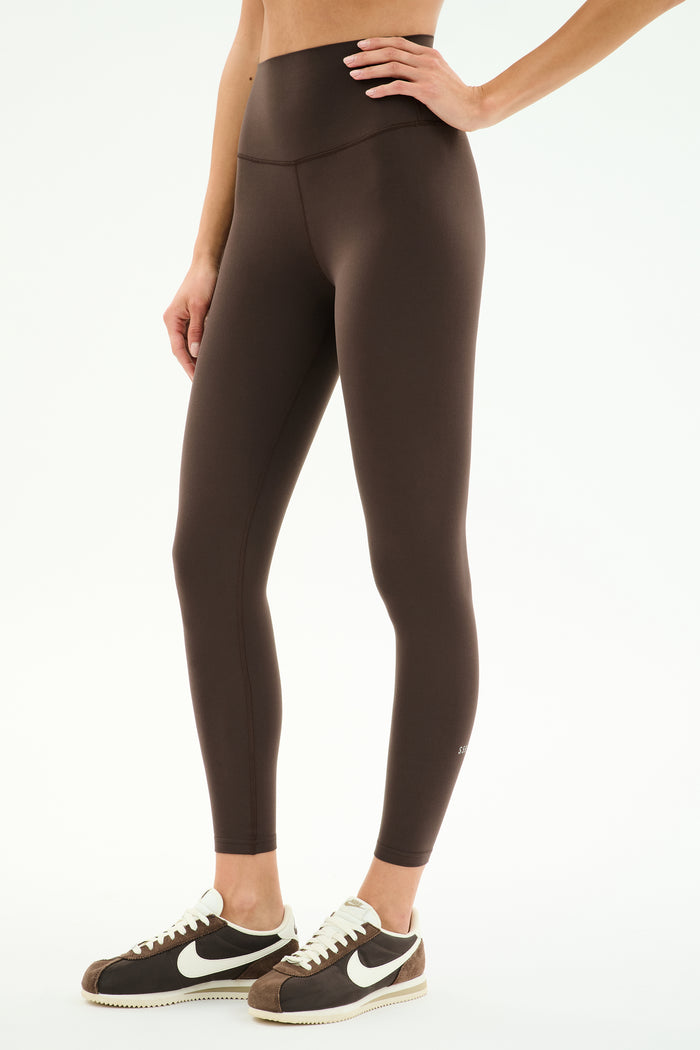 A person wearing SPLITS59 Airweight High Waist Legging in Dark Chocolate and black and white Nike sneakers stands against a plain white background.