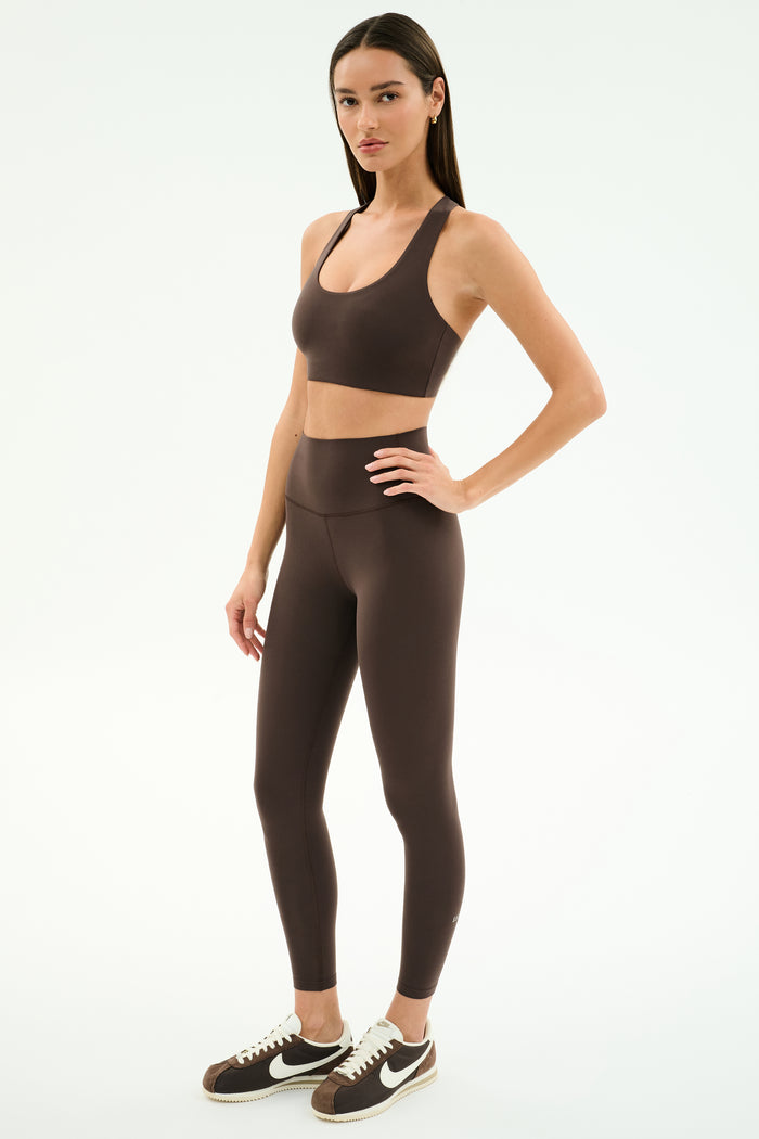A woman stands in a studio against a white background wearing SPLITS59's Airweight High Waist Legging in Dark Chocolate with a matching brown sports bra and brown Nike sneakers—ideal for hot yoga.