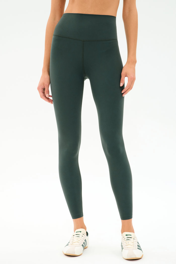 A person stands against a plain white background wearing SPLITS59 Airweight Rib High Waist 7/8 leggings in Military and white sneakers.