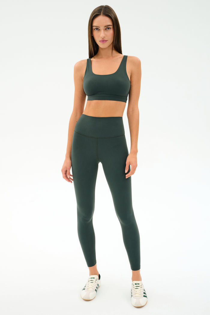 A woman faces forward wearing the SPLITS59 Airweight Rib High Waist 7/8 leggings in Military, paired with a matching sports bra and white sneakers against a plain white background.