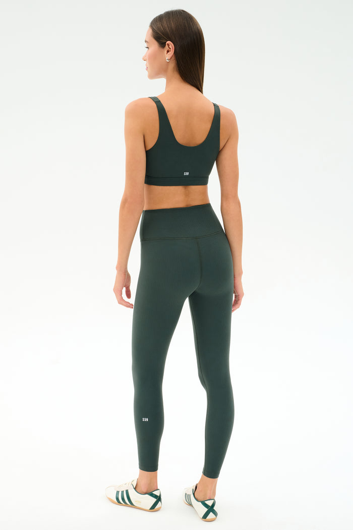 A woman faces away, wearing SPLITS59's Airweight Rib High Waist 7/8 leggings in Military with a matching sports bra and white sneakers, set against a plain white background.
