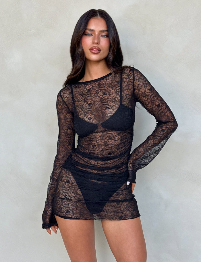 A woman with long dark hair poses confidently in the ALAYNA THREE PIECE MINI DRESS - BLACK: BLACK LACE from TIGERMIST, featuring a sheer black lace material over a black bra and panties. The dress has long sleeves and fits snugly. She stands against a neutral-colored background, her expression poised. The overall look is stylish and bold.