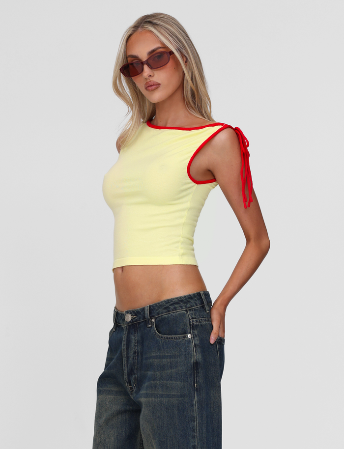 A woman with long blonde hair and sunglasses wears high-waisted blue jeans and the TIGERMIST ALIRA TOP in yellow, featuring a red contrast neckline and shoulder tie, standing against a plain light gray background.