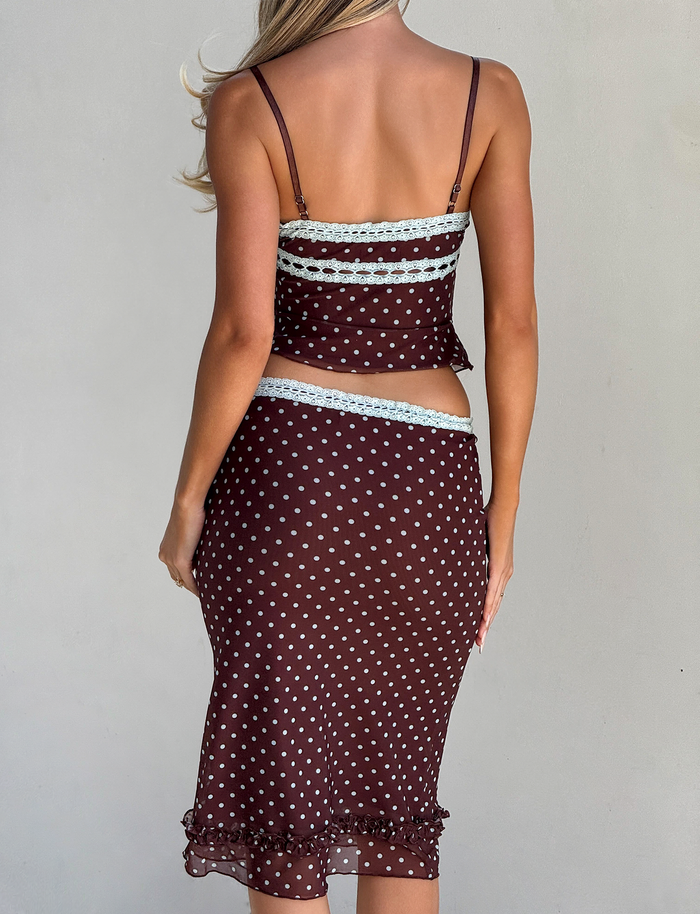 A woman faces away, wearing the TIGERMIST AMARA MIDI SKIRT - POLKA DOT set with brown polka dots, light blue lace accents at the top and waist, and a mesh midi skirt featuring a ruffled hem.