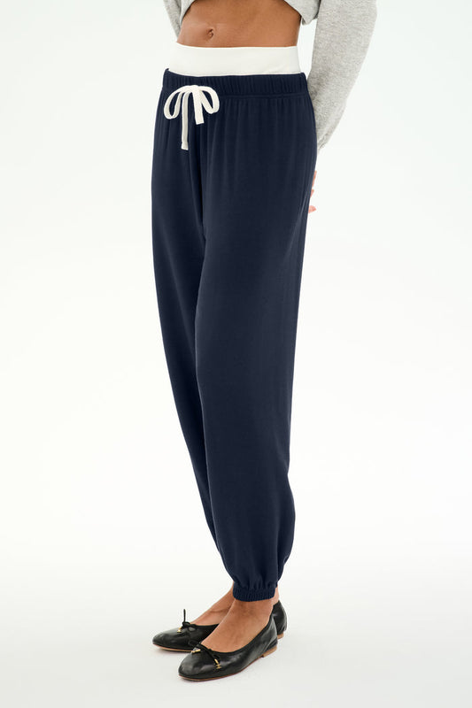 A person is wearing the SPLITS59 Andie Dual Waist Sweatpant in Indigo/White, featuring a comfortable baggy sweatpant fit and a white drawstring. The joggers are styled with a gray sweater and black ballet flats, against a white background. Made in Los Angeles.
