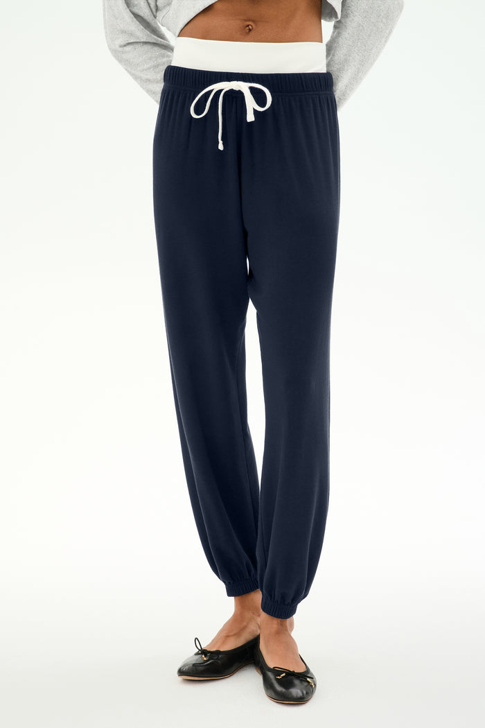 A person wears the SPLITS59 Andie Dual Waist Sweatpant in Indigo/White, featuring a relaxed fit with a white drawstring, paired with a gray top and black ballet flats, standing against a white background. These fleece sweatpants are crafted in Los Angeles to offer effortless comfort and style.