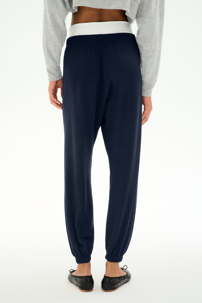 A person stands facing away against a plain white background, wearing the SPLITS59 Andie Dual Waist Sweatpant in Indigo/White, styled with a gray sweatshirt and black flats. These relaxed sweatpants are made in Los Angeles.