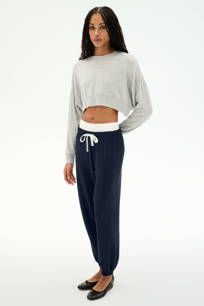 A person stands against a plain white background wearing a light gray cropped sweatshirt, SPLITS59 Andie Dual Waist Sweatpant in Indigo/White (made in Los Angeles), and black flats.