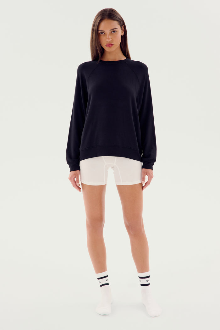 A person standing against a plain background, wearing an SPLITS59 Andie Fleece Sweatshirt - Black made in Los Angeles from stretch modal fabric, white shorts, white socks with black stripes, and white shoes.
