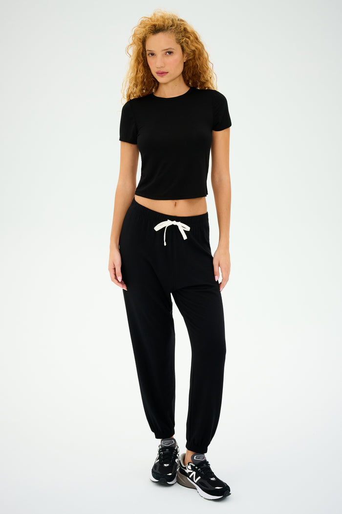 With curly hair, a person wears SPLITS59's Andie Oversized Fleece Sweatpant in black and a simple black t-shirt, matched with sleek sneakers against a plain backdrop. This stylish outfit, ideal for cool weather workouts, is proudly made in Los Angeles.