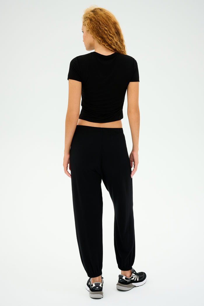 A curly-haired person in a black t-shirt, wearing SPLITS59's Andie Oversized Fleece Sweatpants and black sneakers, stands against a light gray background. Ideal for cool weather workouts.