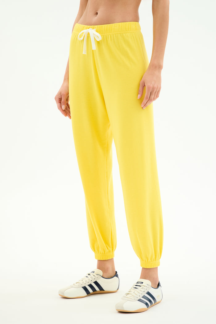 A person wears the SPLITS59 Andie Oversized Fleece Sweatpant in Bright Yellow, featuring a soft spandex blend and white drawstring, paired with white sneakers with dark stripes, standing against a plain white background.