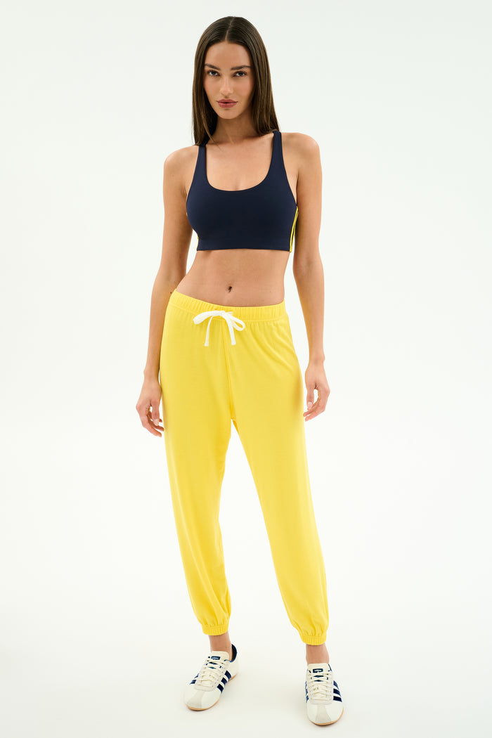 A woman stands against a white background wearing navy spandex blend sports bra, SPLITS59 Andie Oversized Fleece Sweatpant in bright yellow, and white sneakers with blue stripes.