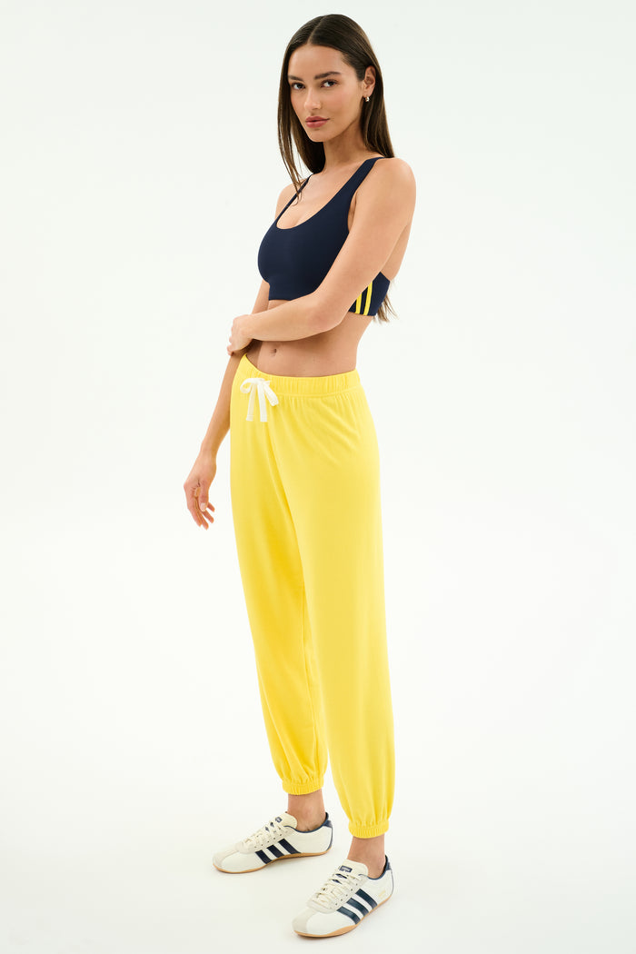 A woman stands in a studio wearing a navy spandex blend sports bra, SPLITS59 Andie Oversized Fleece Sweatpant in bright yellow with a white drawstring, and white Adidas sneakers.