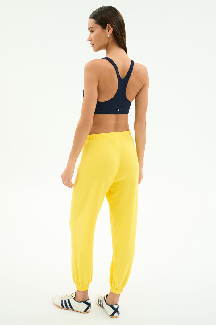 A woman faces away, wearing SPLITS59's Andie Oversized Fleece Sweatpant in bright yellow and a navy sports bra, paired with white athletic shoes with navy stripes, all set against a plain white background.