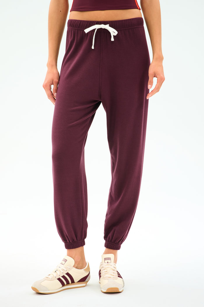 A person wearing the SPLITS59 Andie Oversized Fleece Sweatpant in Dark Cherry, crafted from cozy fleece with a white drawstring, paired with crisp white sneakers featuring burgundy stripes, stands against a plain background—perfect for cool weather workouts.