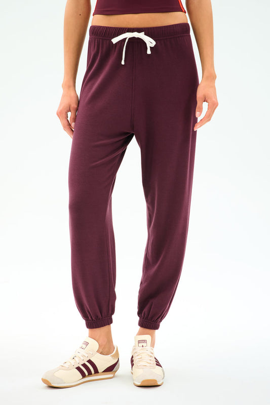 A person wearing the SPLITS59 Andie Oversized Fleece Sweatpant in Dark Cherry, crafted from cozy fleece with a white drawstring, paired with crisp white sneakers featuring burgundy stripes, stands against a plain background—perfect for cool weather workouts.