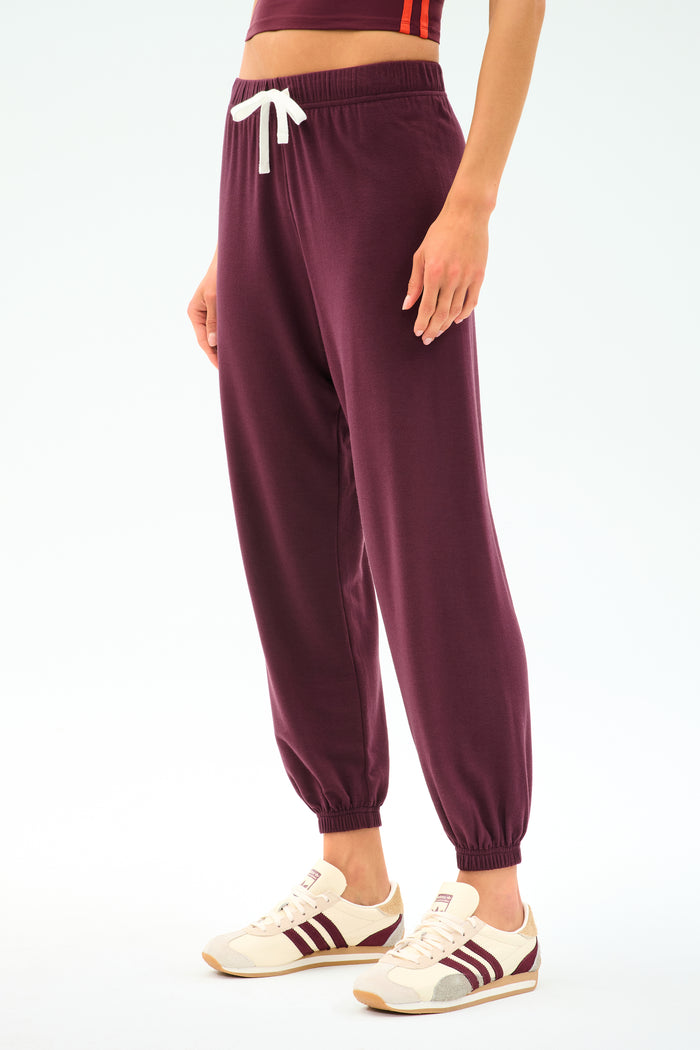 A person models the SPLITS59 Andie Oversized Fleece Sweatpant in Dark Cherry—ultra-luxe, featuring a white drawstring—paired with cream sneakers accented by maroon stripes, ideal for cool weather workouts, set against a plain white backdrop.