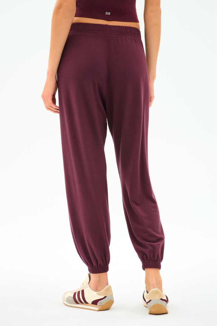 Model wears the SPLITS59 Andie Oversized Fleece Sweatpant in Dark Cherry, paired with beige sneakers featuring maroon stripes. Shown from the back against a white background—ideal for cool weather workouts or relaxing in comfort and style.