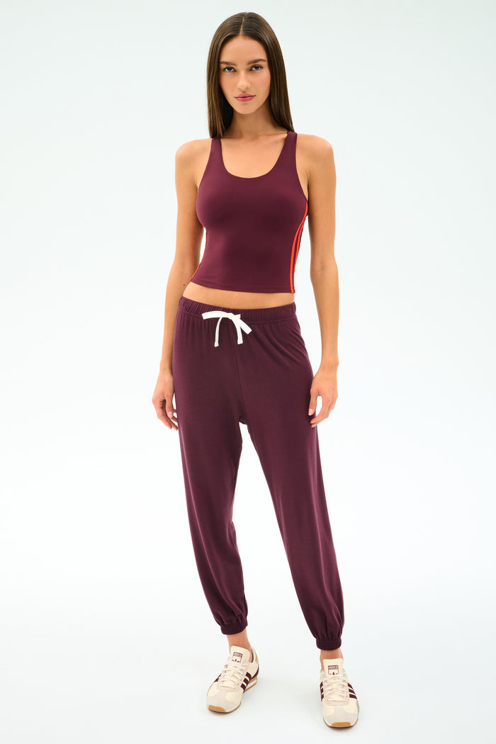 A woman stands against a plain white background wearing the SPLITS59 Andie Oversized Fleece Sweatpant in Dark Cherry, styled with a maroon tank top and beige sneakers—the ultra-luxe jogger features an oversized fit and white drawstring, perfect for cool weather workouts.