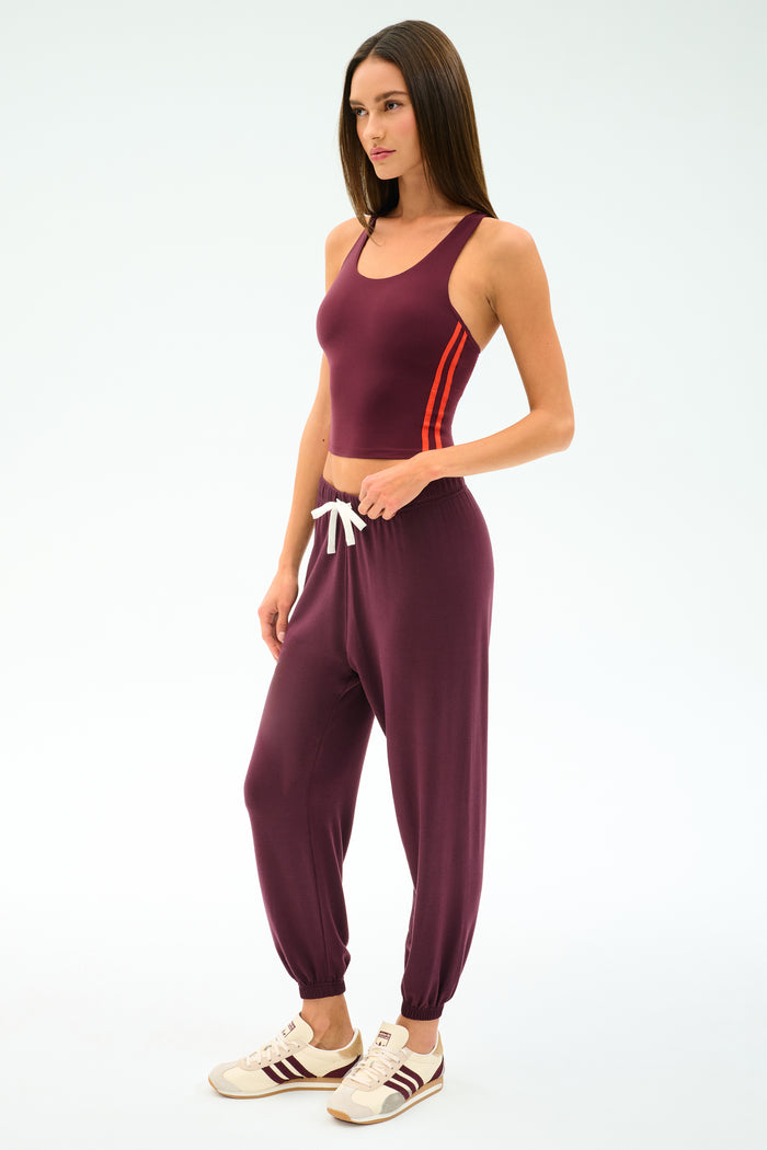 A woman stands against a plain light background wearing the SPLITS59 Andie Oversized Fleece Sweatpant in Dark Cherry, paired with a maroon sleeveless top featuring red side stripes and white and maroon sneakers.