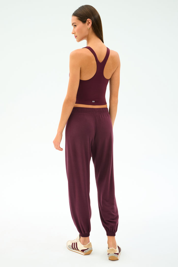 A woman stands facing away, wearing the SPLITS59 Andie Oversized Fleece Sweatpant in Dark Cherry with a matching burgundy racerback sports top and beige and burgundy sneakers, set against a plain light background—perfect for cool weather workouts.
