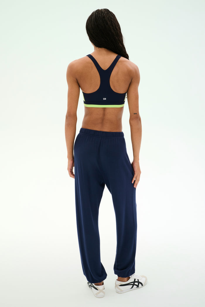 A person stands facing away against a plain light background, wearing a navy sports bra with green trim, white athletic shoes, and the SPLITS59 Andie Oversized Fleece Sweatpant in Indigo—an ideal look for cool weather workouts.