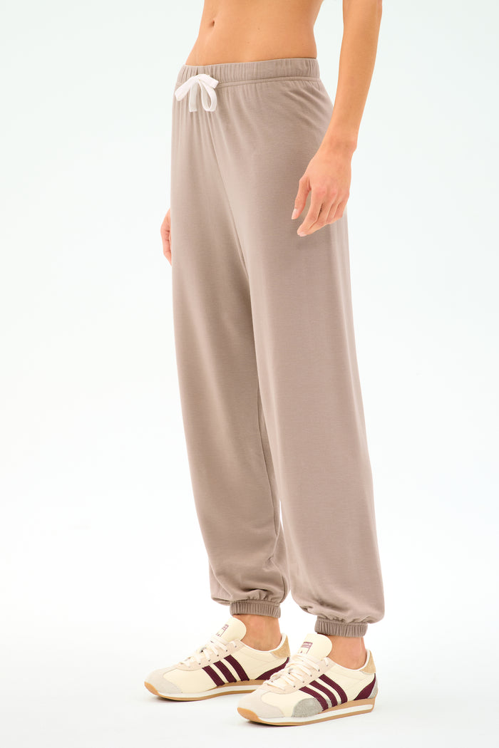 Person wearing the SPLITS59 Andie Oversized Fleece Sweatpant in Latte, featuring a white drawstring and paired with beige sneakers with maroon stripes, perfect for cool-weather workouts against a plain light background.