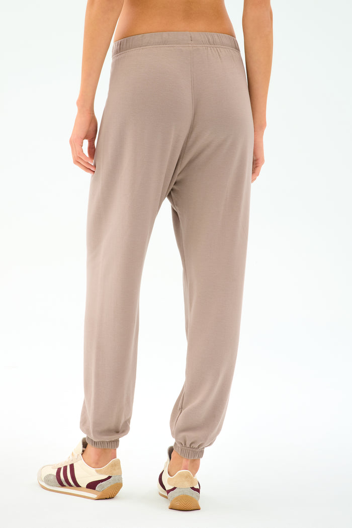 A person stands against a plain white background wearing SPLITS59’s Andie Oversized Fleece Sweatpant in Latte and white sneakers with maroon accents, an ideal look for cool-weather workouts.