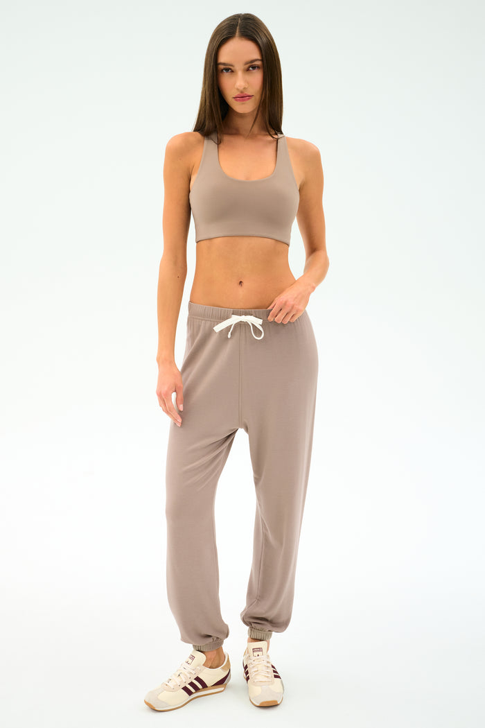 A woman stands against a plain background wearing a beige sports bra, SPLITS59 Andie Oversized Fleece Sweatpant in Latte with a drawstring, and white sneakers with maroon stripes—ideal for warm-ups or cool weather workouts.