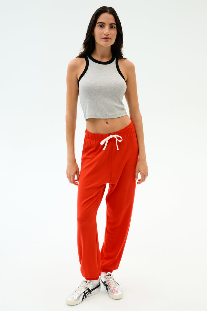 A woman faces forward in a gray sleeveless crop top and SPLITS59 Andie Oversized Fleece Sweatpant in Lotus Red with a white drawstring, paired with white sneakers, set against a plain white background.