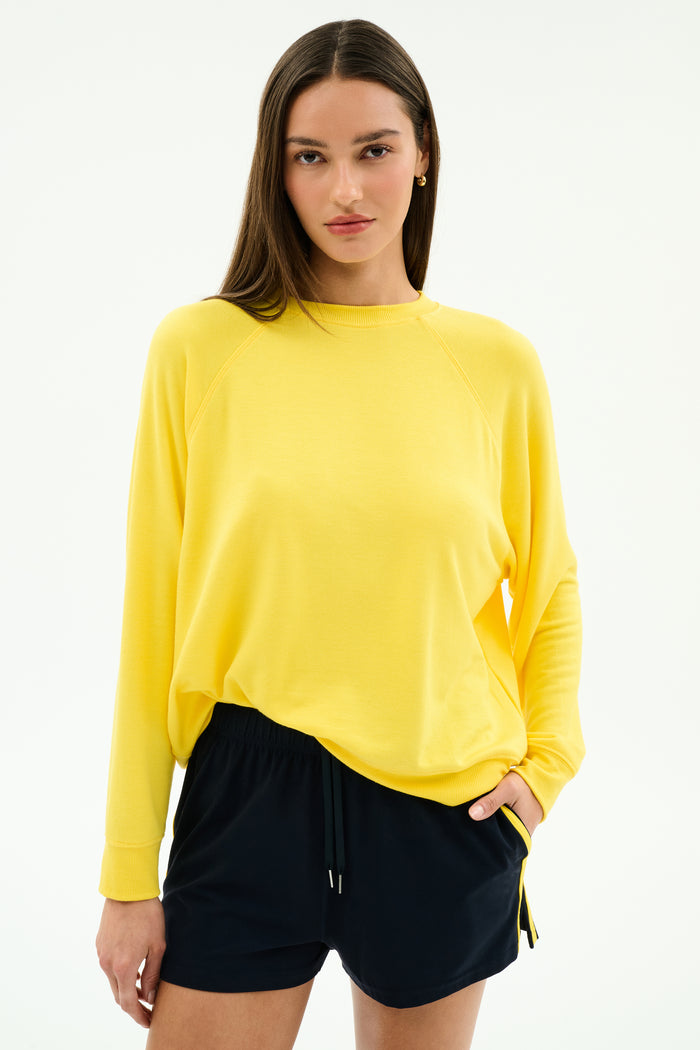 A woman stands against a plain background wearing the SPLITS59 Andie Oversized Fleece Sweatshirt in Bright Yellow, paired with black drawstring shorts, with one hand in her pocket.