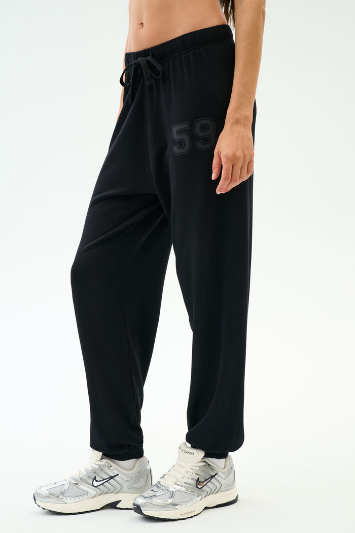 A person wears the SPLITS59 Andie Oversized Fleece Varsity 59 Sweatpant - Black/59, featuring a "59" print on the left thigh, paired with white sneakers and a crop top—perfect for cool weather workouts—against a white background.