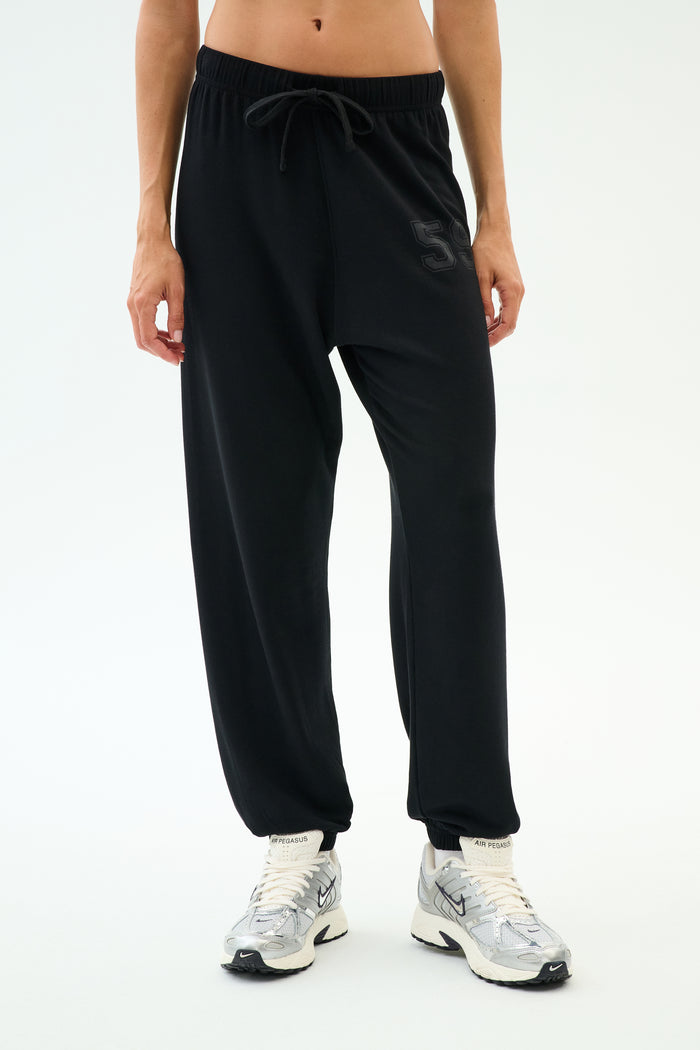 A person wears SPLITS59's Andie Oversized Fleece Varsity 59 Sweatpant in black, featuring a "59" on the left thigh, paired with white sneakers—ideal for cool weather workouts—standing against a plain white background.