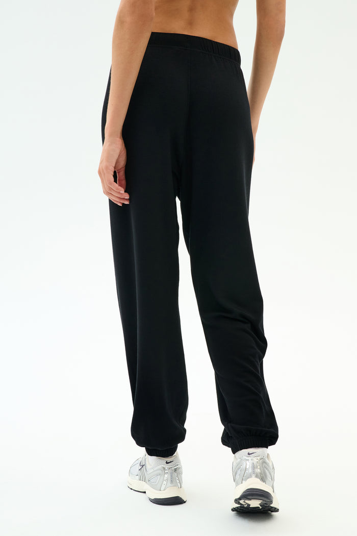 A person wears SPLITS59’s Andie Oversized Fleece Varsity 59 Sweatpant in Black/59 with white sneakers, standing on a white background and facing away from the camera—perfect for cool weather workouts.