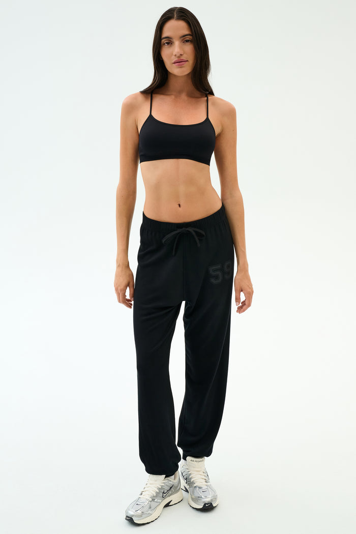 A woman stands against a plain background in SPLITS59’s Andie Oversized Fleece Varsity 59 Sweatpant in Black, paired with a black sports bra and white sneakers—perfect fleece joggers for cool weather workouts. Made in Los Angeles.