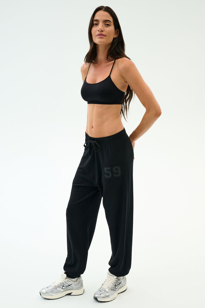 A woman stands in a studio wearing a black sports bra, white sneakers, and SPLITS59’s Andie Oversized Fleece Varsity 59 Sweatpant in Black/59—ultra luxe fleece with "59" on the leg—perfect for cool weather workouts.