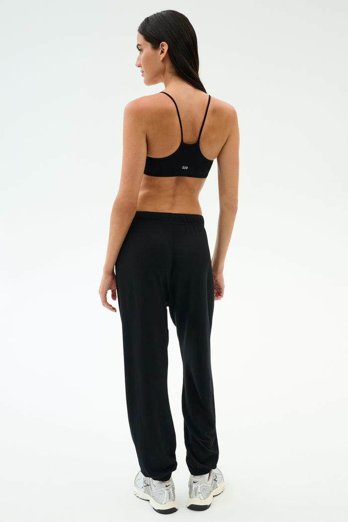 A woman stands with her back to the camera in SPLITS59’s Andie Oversized Fleece Varsity 59 Sweatpant - Black/59, paired with a black sports bra and white sneakers, ideal for cool weather workouts against a plain light background.