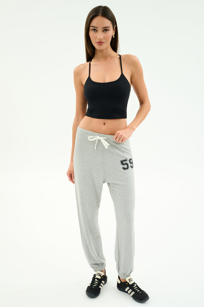 A woman stands against a plain background wearing a black tank top, SPLITS59 Andie Oversized Fleece Varsity 59 Sweatpant in Heather Grey with "59" on the hip, and black sneakers.