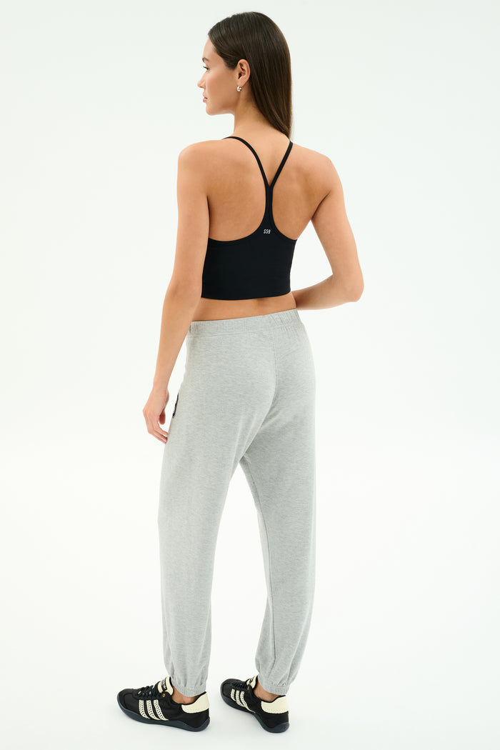 A woman faces away wearing a black sports bra, SPLITS59 Andie Oversized Fleece Varsity 59 Sweatpant in Heather Grey/59, and black athletic shoes with white stripes against a plain white background.
